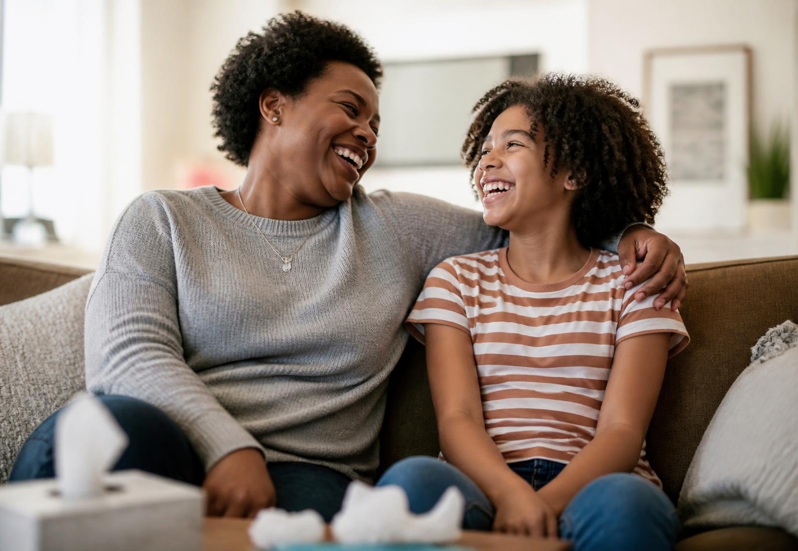 A caregiver and a child laughing together on the couch after a hard moment, arm around her shoulders.