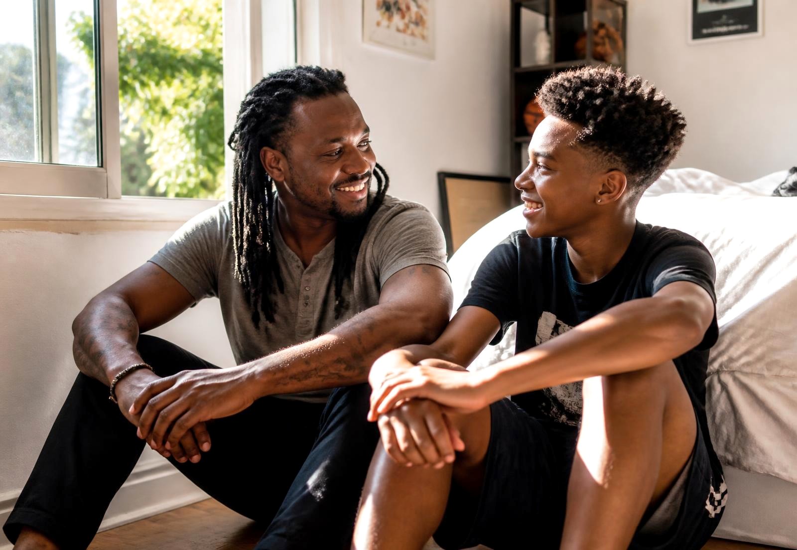 A father with locs and his teenage son sitting on the floor by a bright window, smiling as they talk.