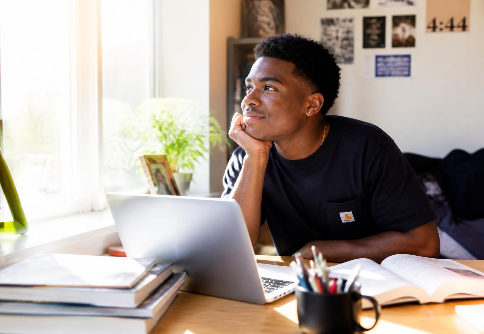 A college student at a sunlit desk full of books, chin in hand, thoughtful and hopeful as light streams through the window.